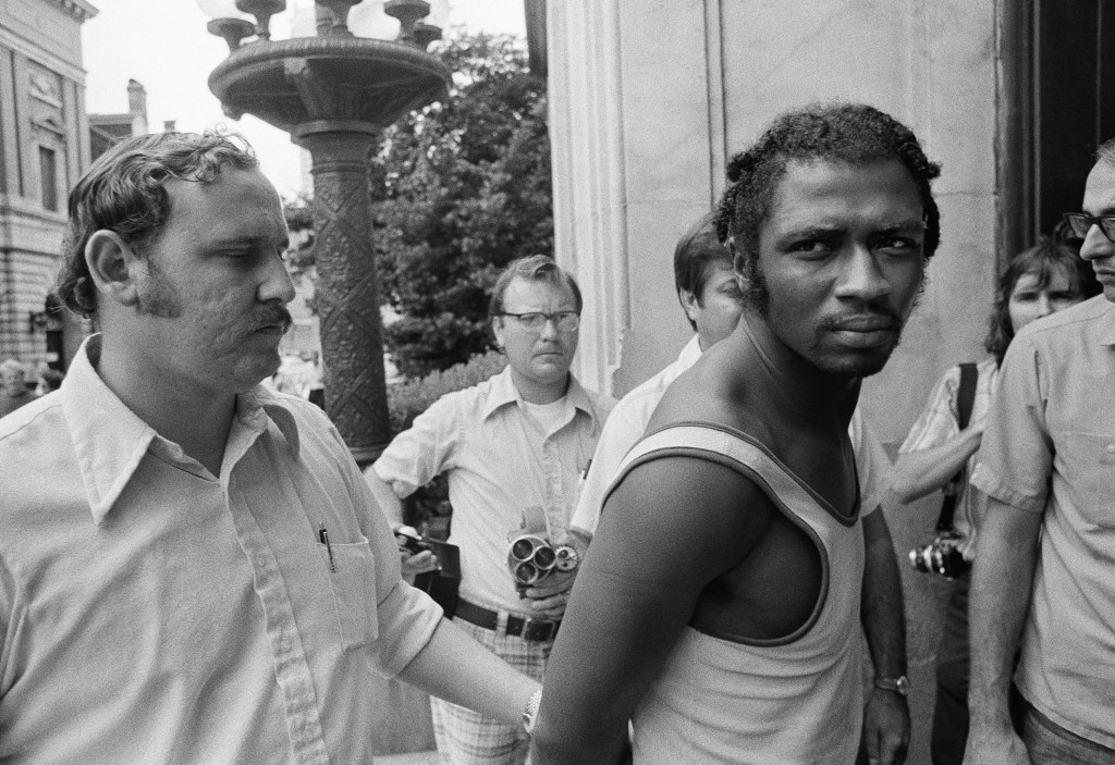 black and white image of herman bell in a white tank top with his arms cuffed behind his back looking at the camera.
