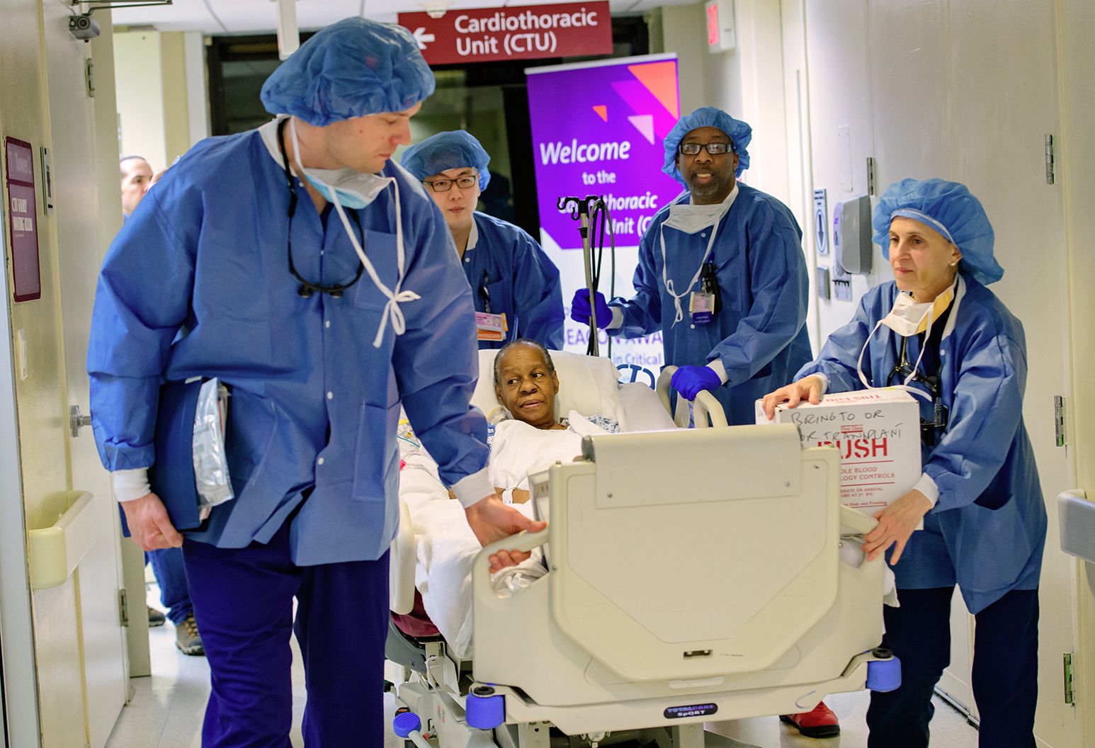 An elderly woman is lying in a hospital bed. Her medical team of 4 people dressed in blue surgical scrubs is wheeling her through the hallway of a hospital.